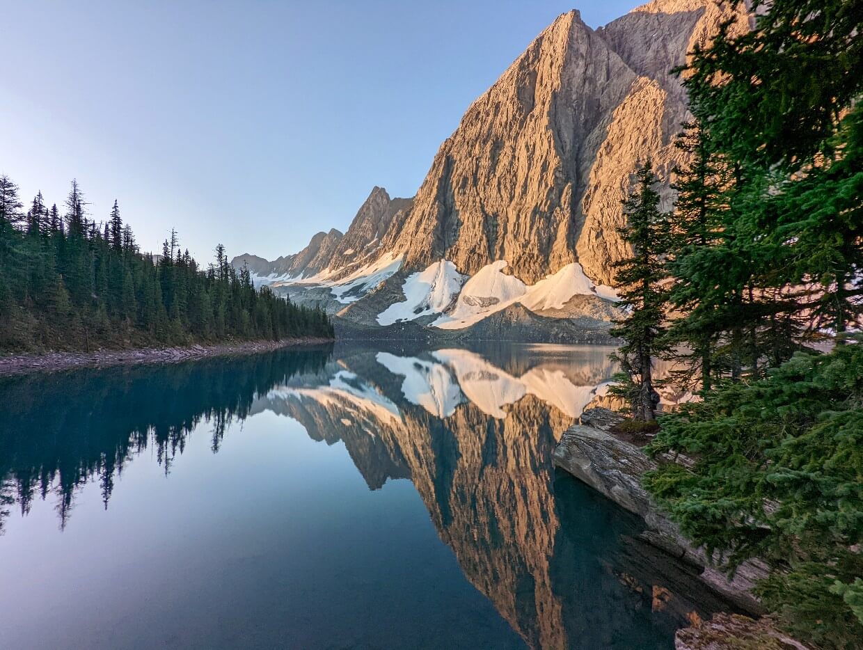 Perfectly calm Floe Lake with reflections of tall mountains above and snow piles on the base