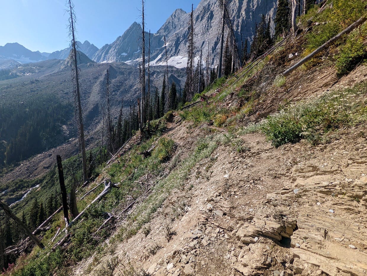 A narrow dirt path curves around the side of a slope, with mountains visible in the background