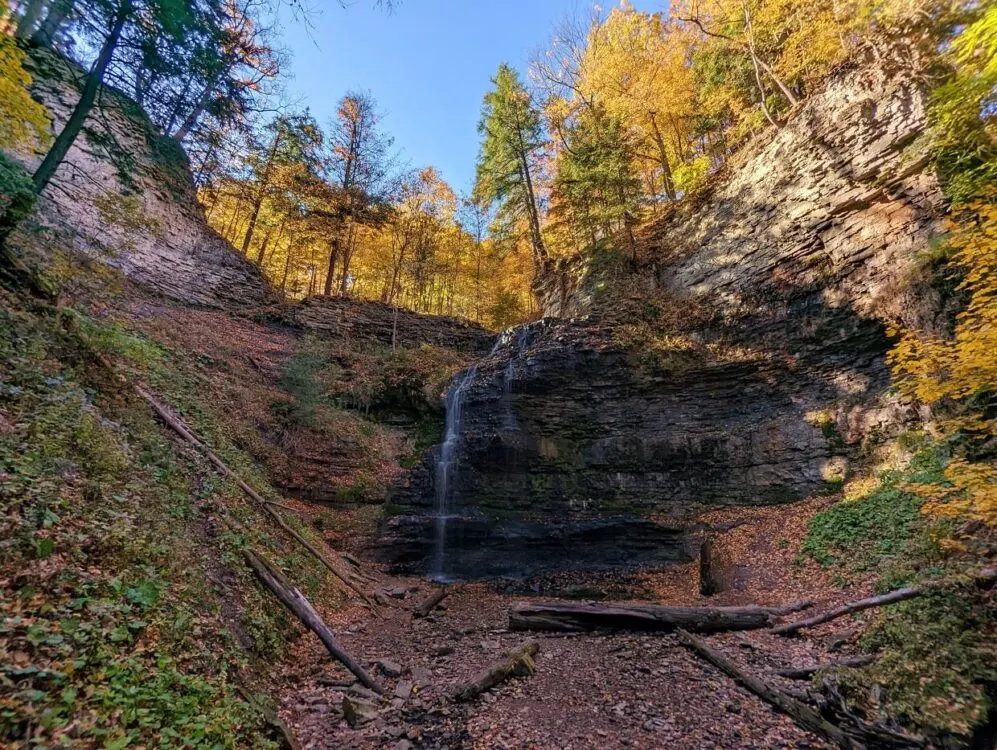 Looking across to cascading waterfall in a rock bowl, with multiple streams of water. The trees in the background are yellow