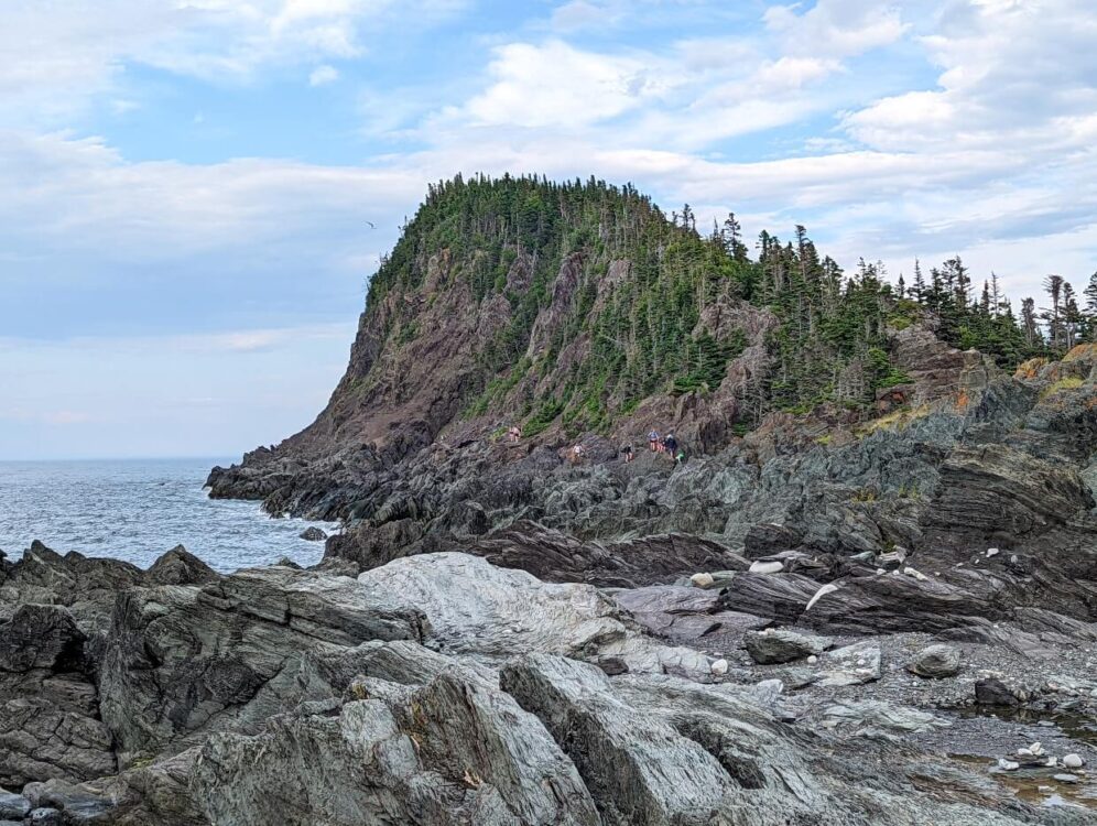 Looking across jagged rocks to rocky hiking trail, with colourful hikers picking their way along the coastline