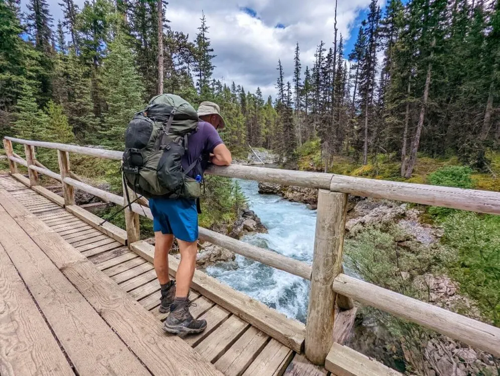 Back view of JR standing on bridge with large backpack, looking down on rushing river on Mount Shark route to Mount Assiniboine