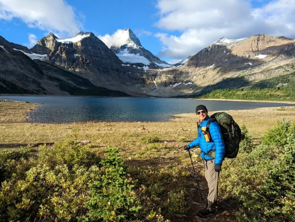 JR stands in front of Magog Lake in blue jacket, carrying large green backpack. The lake is calm and the peak of Mount Assiniboine can be seen behind. JR has a bright orange bear spray holster on his shoulder strap