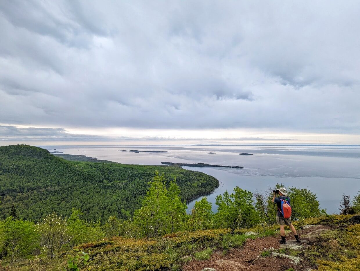 Back view of JR in hiking attire, taking a photo of something in the distance, with views of forest and lake below