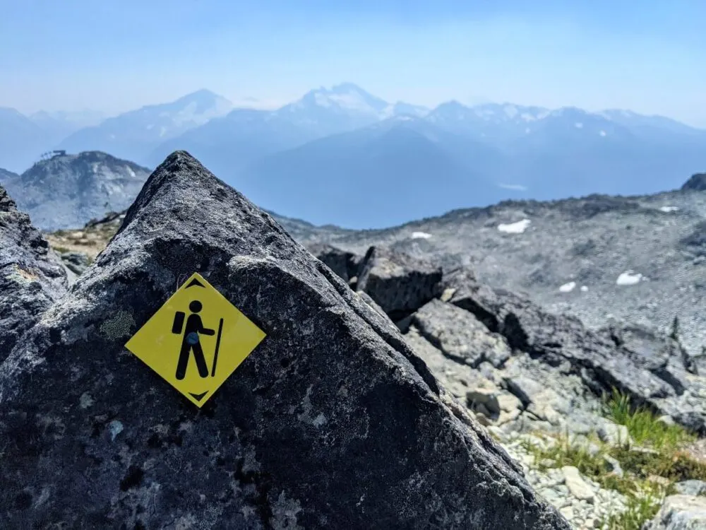 Yellow hiker sign on rock at Whistler Mountain, with snow capped peaks in background