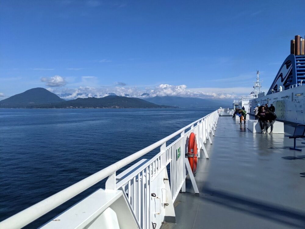 Standing aboard BC Ferries outside deck, with calm ocean and islands visible on left and boat funnels on right