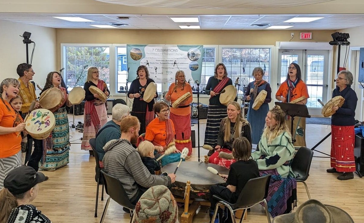 Boundary Métis Drummers drum circle at Christina Lake Winterfest, with additional drummers lined up behind. A number of the drummers are wearing orange t-shirts as a symbol of the forced assimilation of Indigenous children in Canadian residential schools