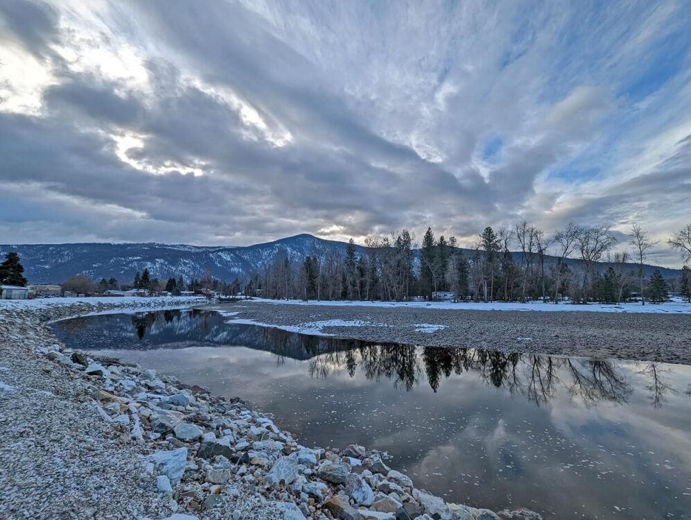 Shore view of Kettle River in Grand Forks, with reflective water and trees and snowy mountains in background