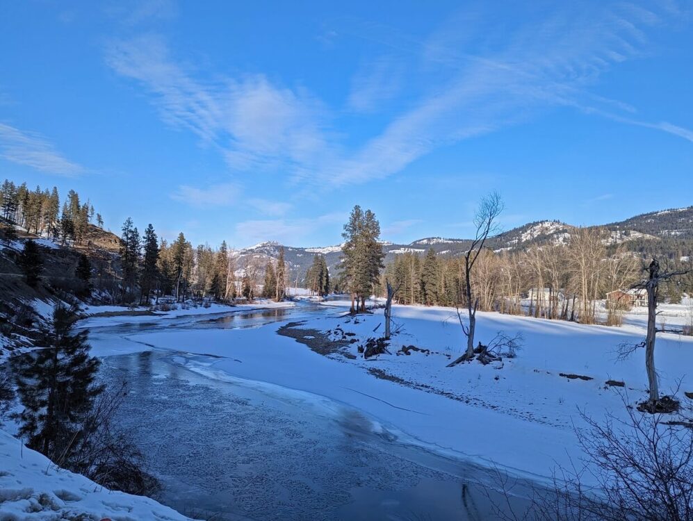 Looking across frozen Kettle River in Rock Creek, with forest and mountains in background