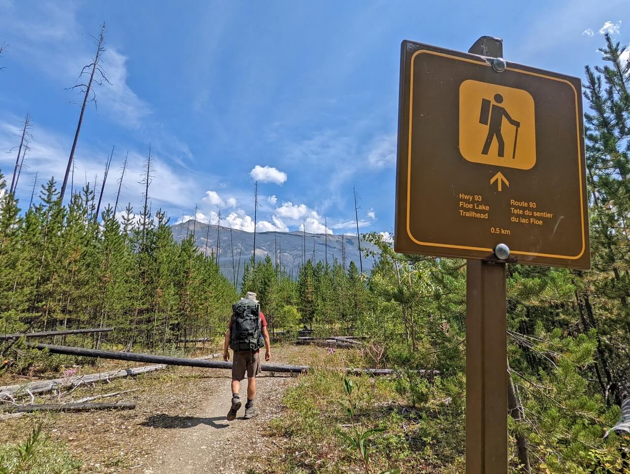 Back view of JR hiking past brown metal Highway 93 parking sign on the Floe Lake Trail