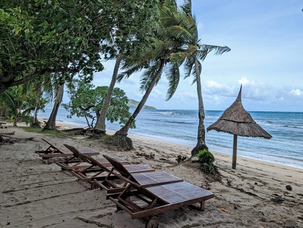 Wooden beach loungers on sandy Blue Lagoon Resort beach. The palm trees above are being blown by strong winds. Large waves are visible on the ocean