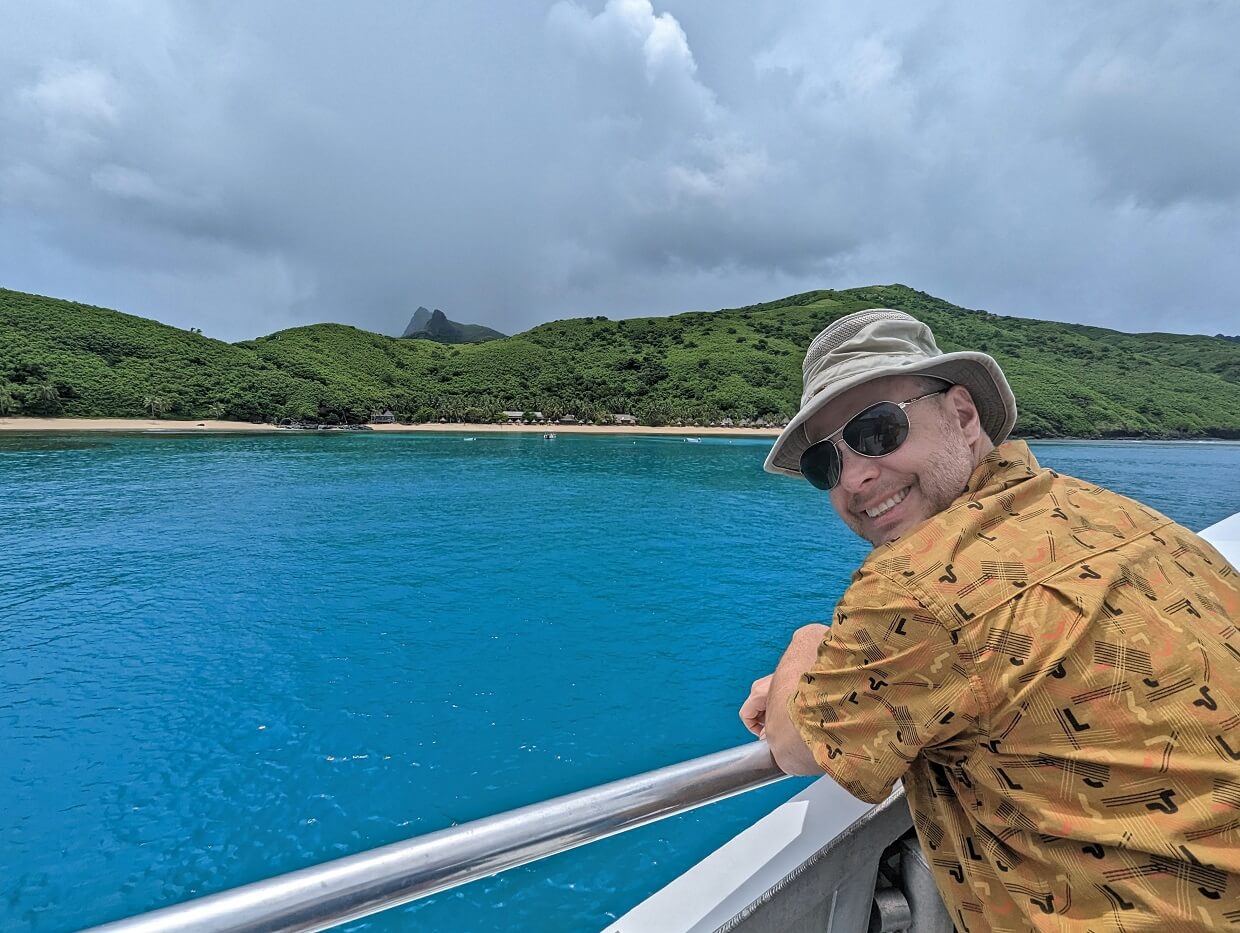JR standing on boat looking back to camera, in front of deep aquamarine ocean and beach resort in background. The sky is gray and rain is falling in the hills behind the resort