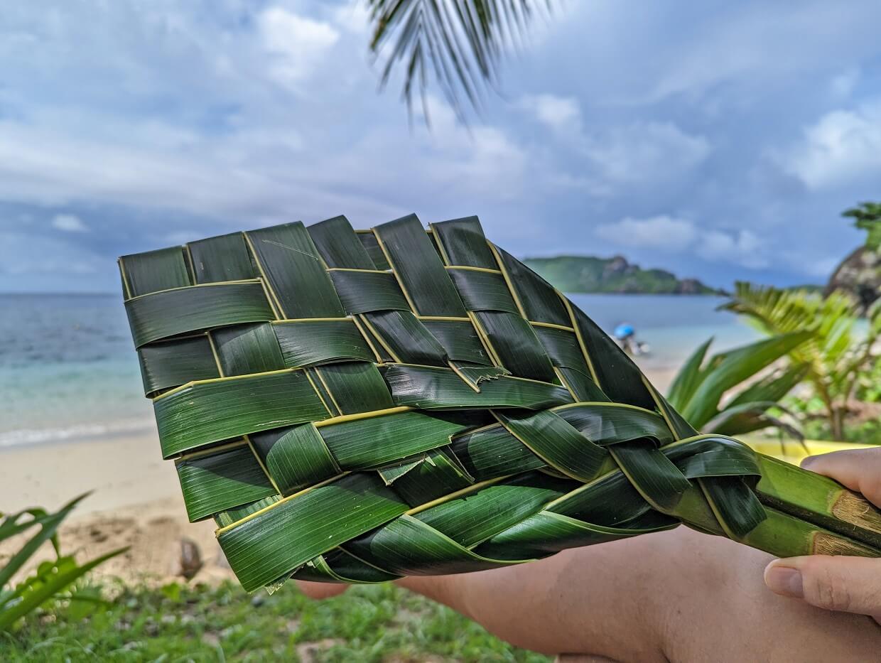 A hand holds handmade palm fan in Fiji, in front of beach view