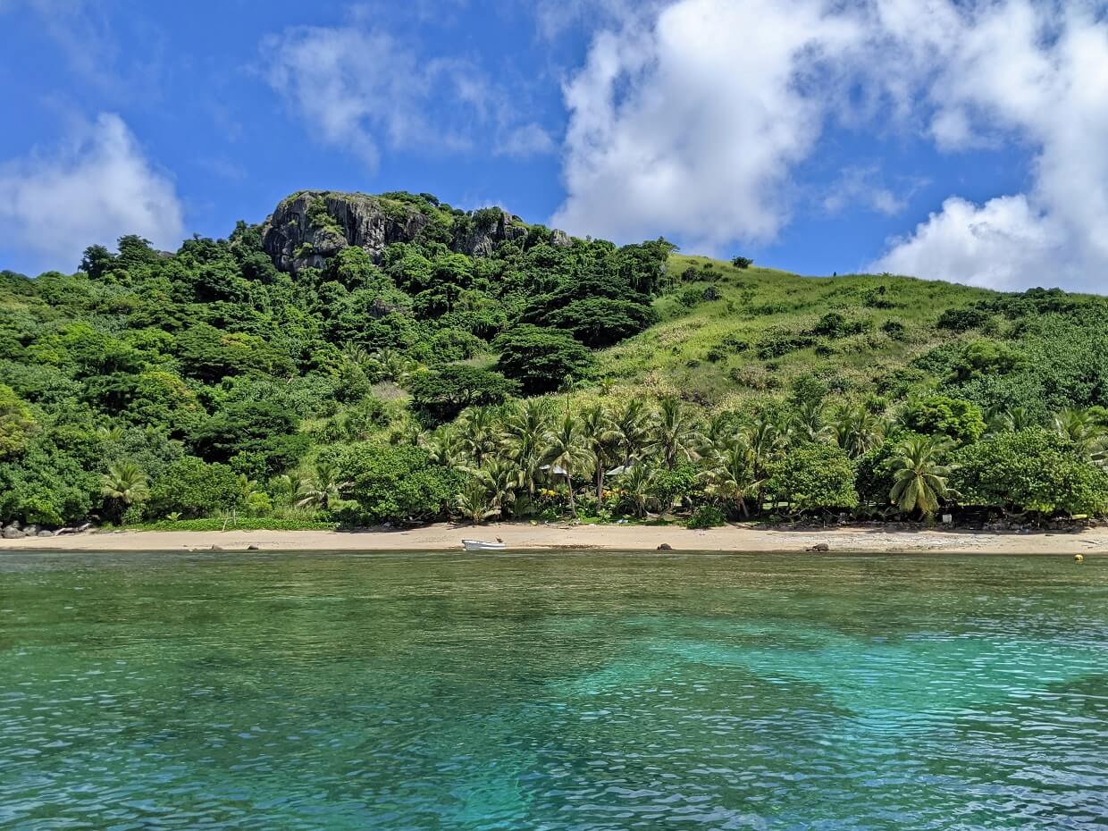 Ocean view of Naqalia Lodge with golden sand beach in distance and volcanic hills above (exposed rock). The ocean is patchy aquamarine, hinting at coral below