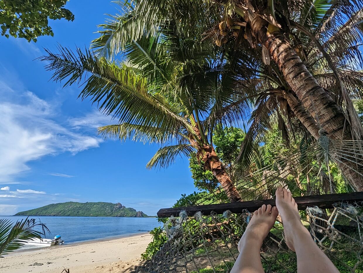 Hammock view looking towards feet and palm trees above, with sandy beach on left and blue skies