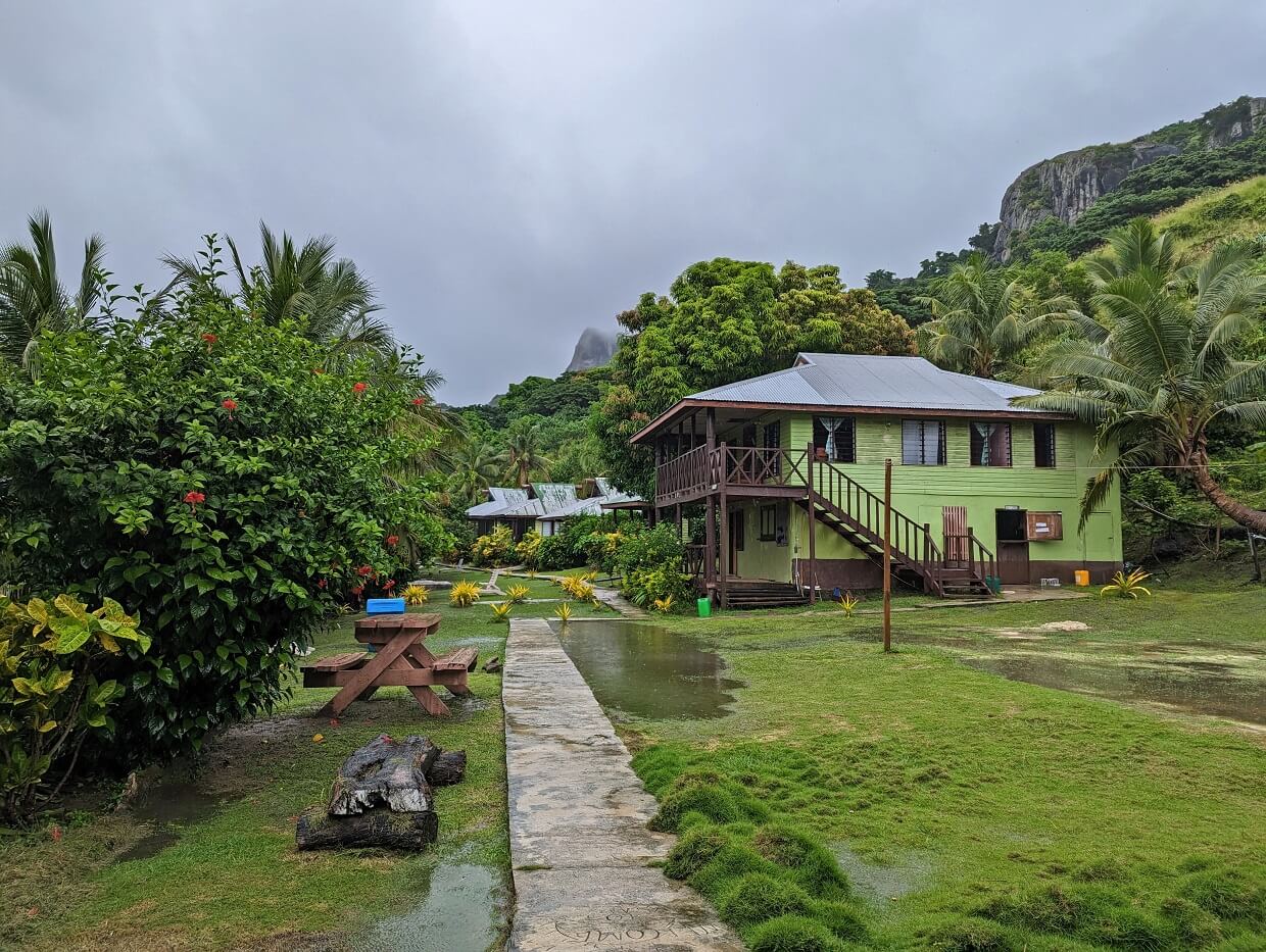 A concrete path leads towards green coloured buildings at Naqalia Lodge, with large rain puddles on surrounding grass. The sky is gray