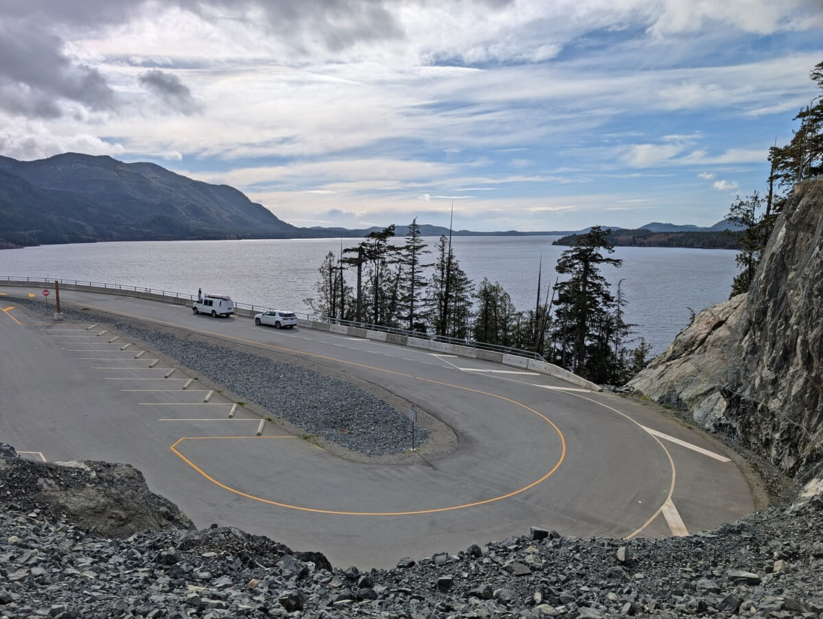 Looking down on Kennedy Lake Lookout, which features a large oval parking area next to the expansive lake. There are two vehicles parked on the edge of the lookout