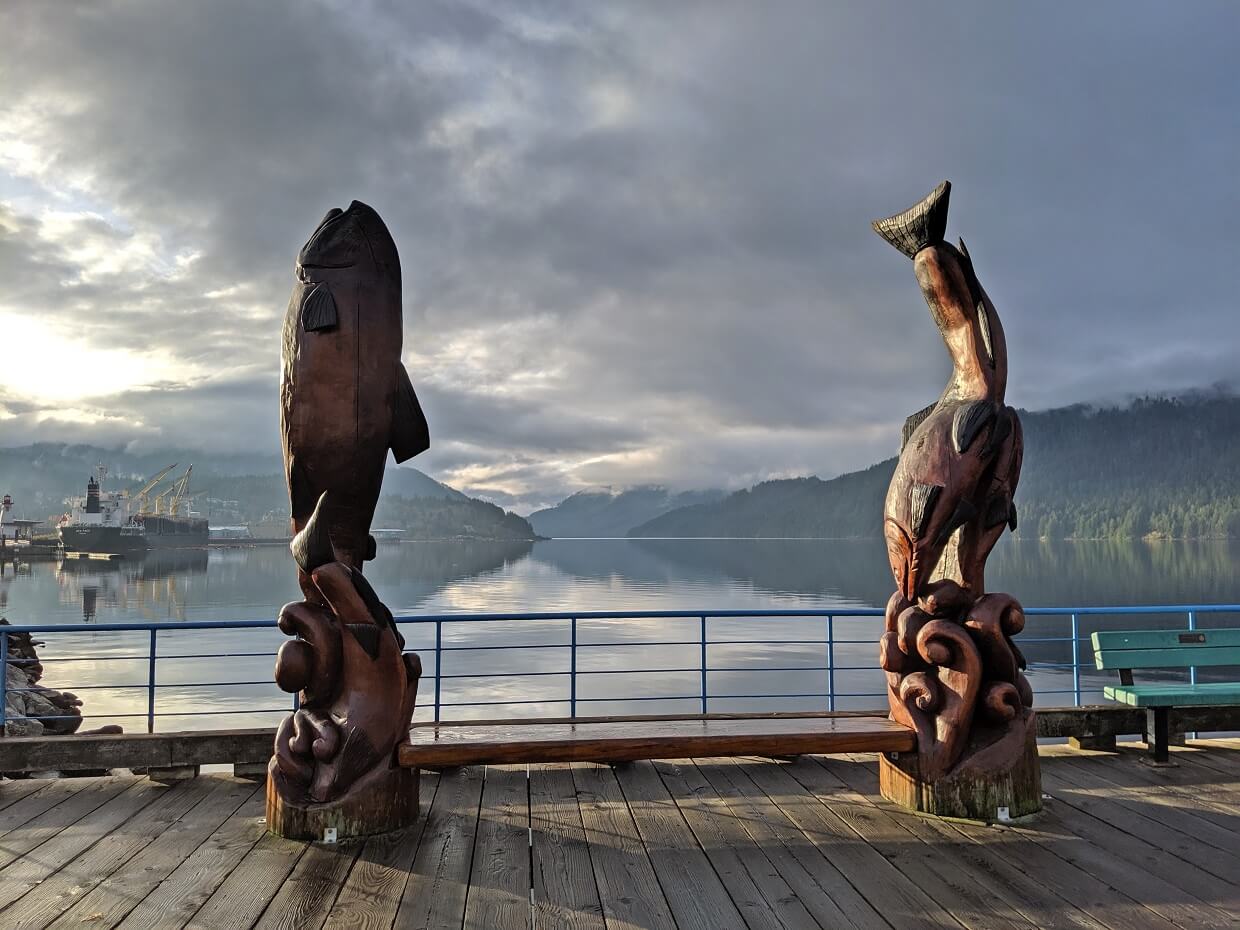 Two wooden sculptures sit on wooden boardwalk path overlooking Alberni inlet in Port Alberni, with reflective ocean water and misty mountains in the background