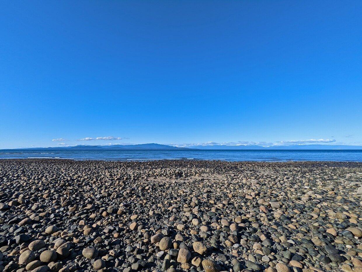 Looking across pebble section of Rathtrevor beach with calm ocean and mountains visible in background