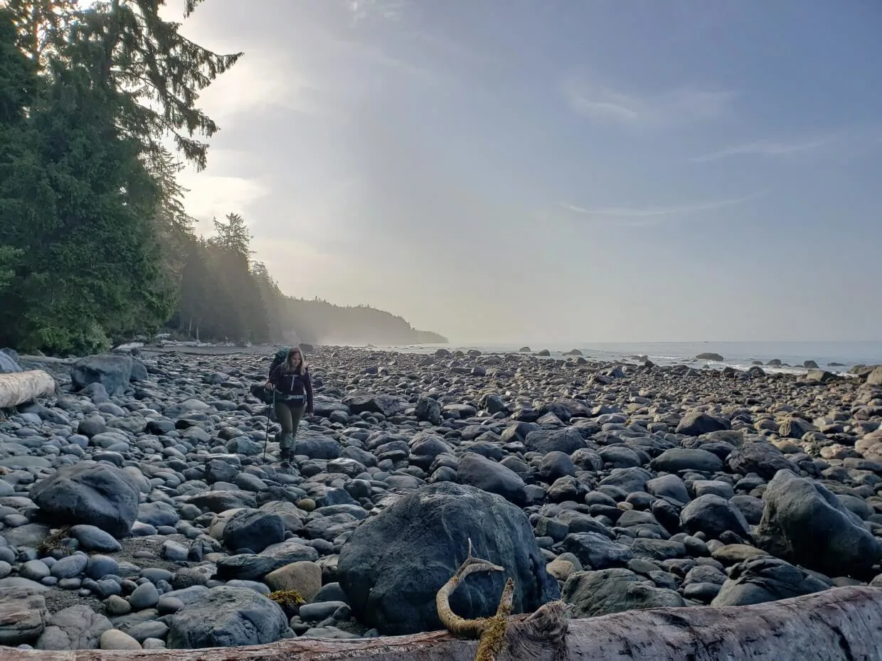 Looking east on rocky coastal beach on the Juan de Fuca Trail, with a hiker walking towards camera. The beach is bordered by forest to the left and ocean to the right