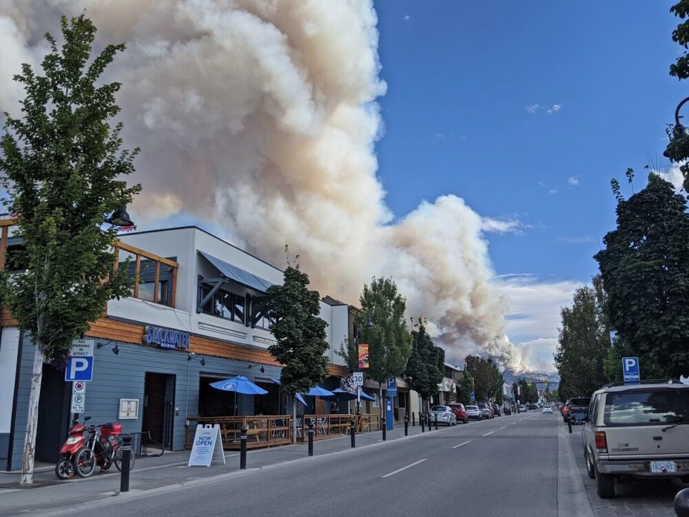 Penticton street view with parked cars on right, brewery on left and wildfire visible in distance, indicated by large plume of smoke
