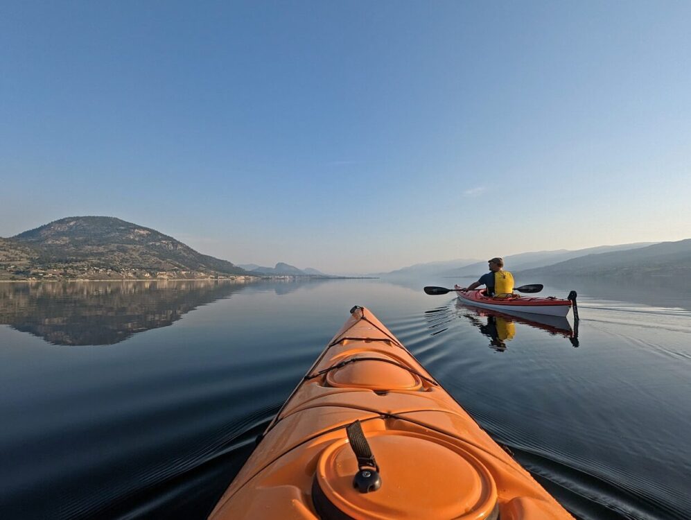 Kayak view of calm, reflective Okanagan Lake, with JR in red kayak to right. There is a large forested mountain on the left hand side. The mountains in the distance are obscured by wildfire smoke