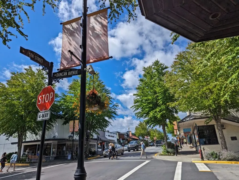 Street scene in dpowntown Courtenay with four way crossing, trees lining sidewalk, road signs and stop sign on left, downtown banners above
