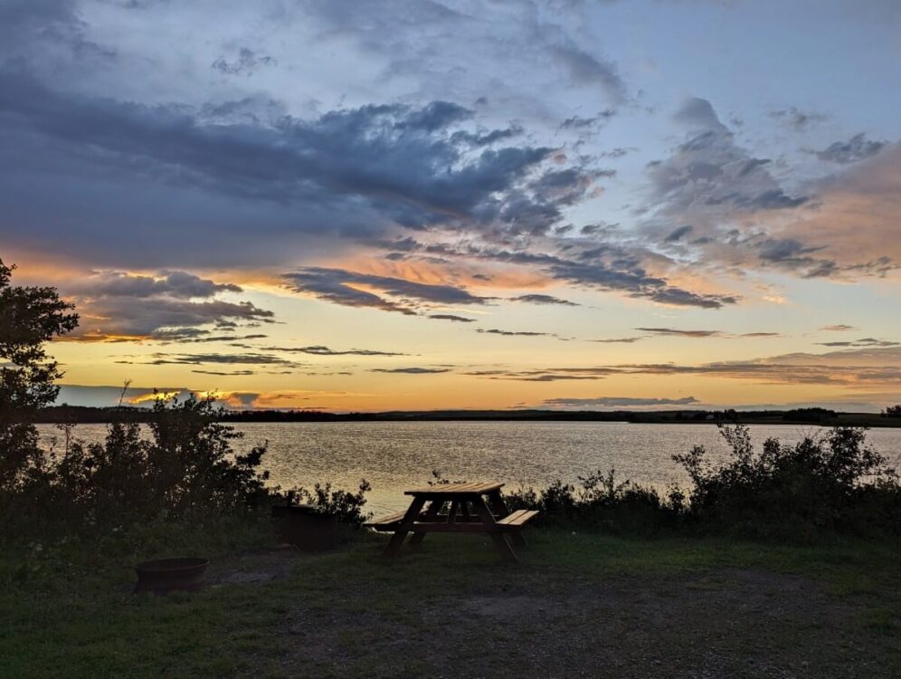 Sunset at Haunted Lakes Campground, with picic table and fire pit visible next to calm lake, with orange and yellow sunset colours above