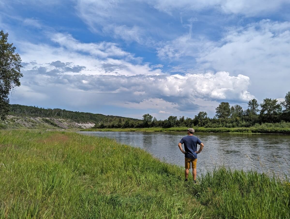 Back view of JR standing in grass next to calm Red Deer River, looking over at clay cliffs in distance