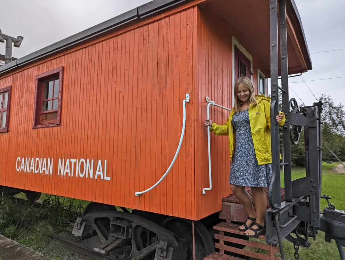 Gemma climbs down steps of bright orange caboose in Mirror, wearing a blue dress and yellow jacket