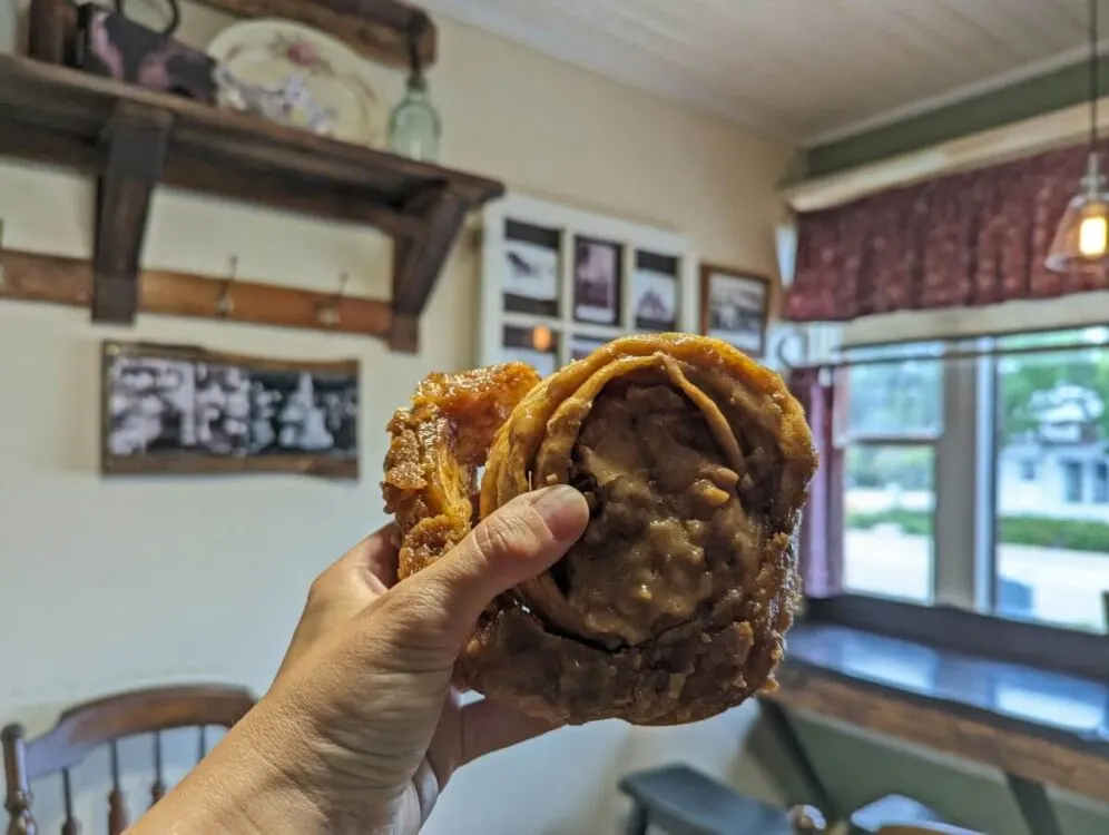 A hand holds up a sticky cinnamon bun in front of the camera, in a cute old fashioned cafe in Alix