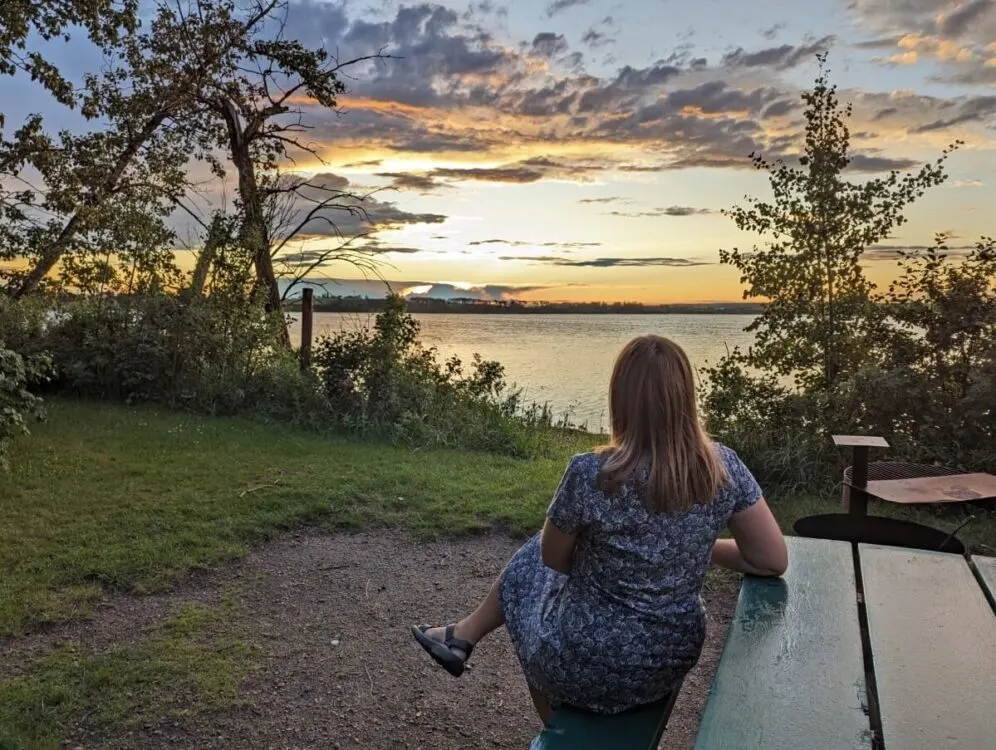 Back view of Gemma sat on picnic bench in front of lake, with bright yellow/orange sunset at horizon