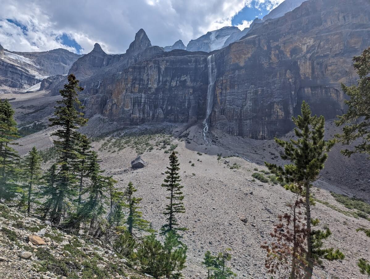 Looking up to a huge waterfall cascading down steep cliffs below Stanley Glacier with boulderfield below, trees in foreground and glacier in background on left