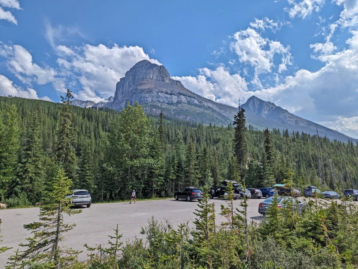Stanley Glacier parking lot, with mountain peaks behind and forest surrounding. There are a dozen vehicles parked in the lot