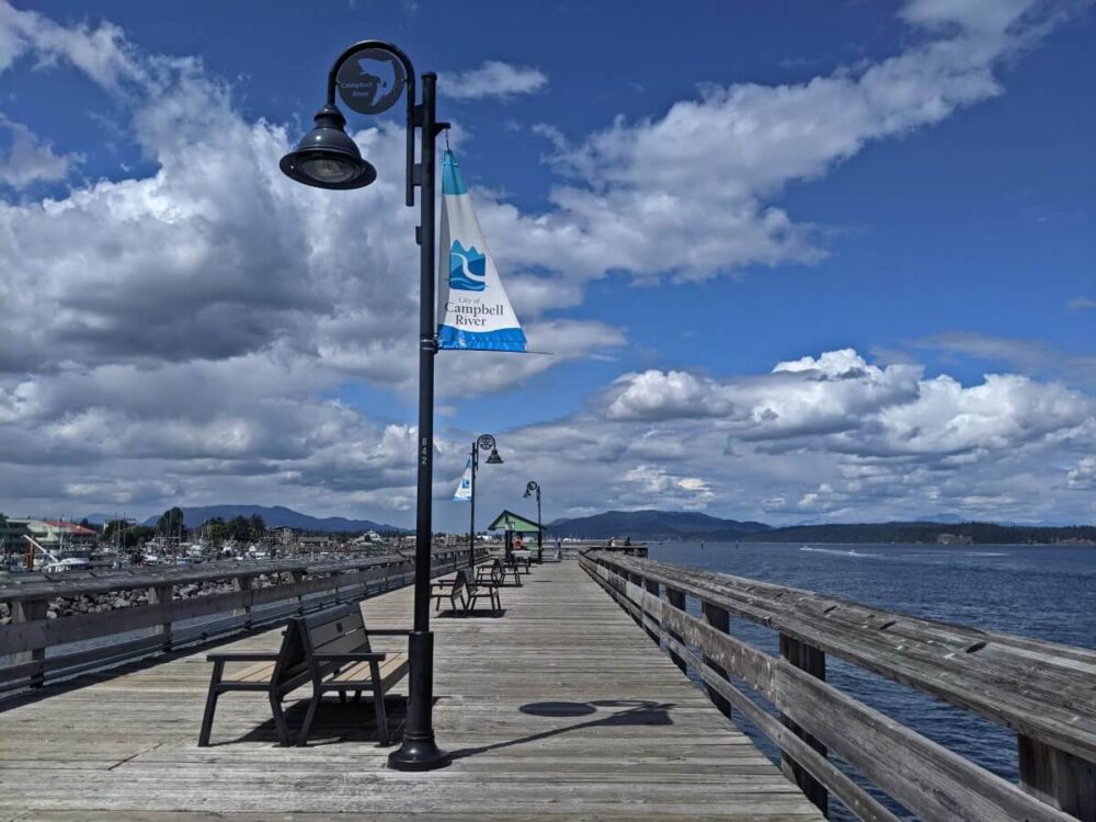 Wooden walking pier in Campbell River, with benches, lightning and signage. The ocean is visible to the right and is calm