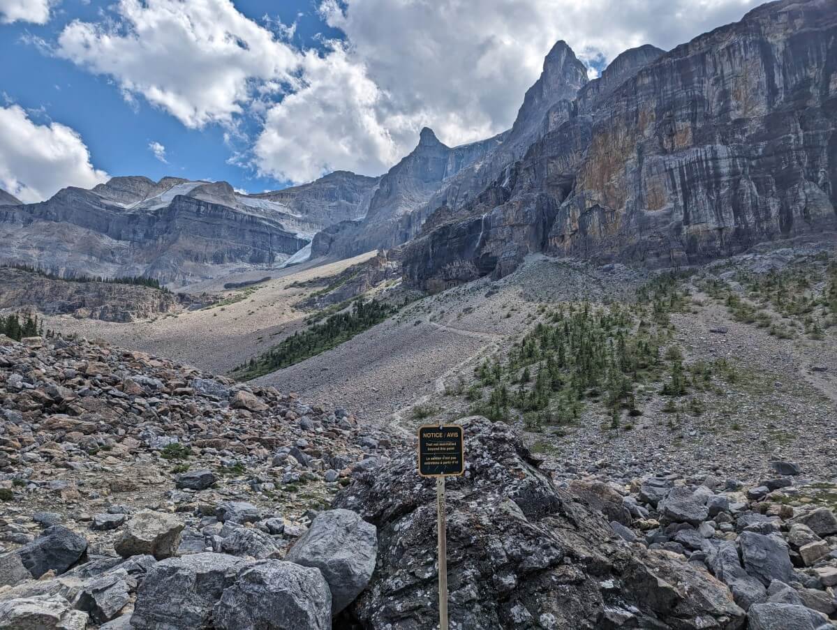 Brown trail end sign in front of large boulders with steep cliffs and glacier in background