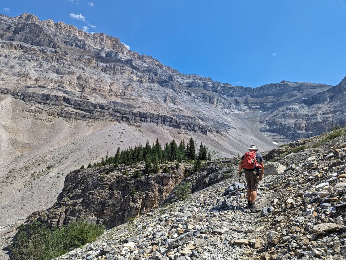 Back view of JR finishing steep scree trail section with forested bench area to left and castle like peaks above