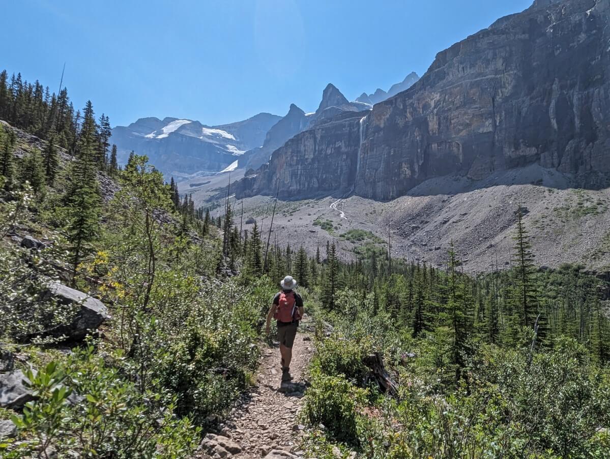 Back view of hiker walking along dirt trail towards mountainous basin area with steep cliffs, waterfall and glacier