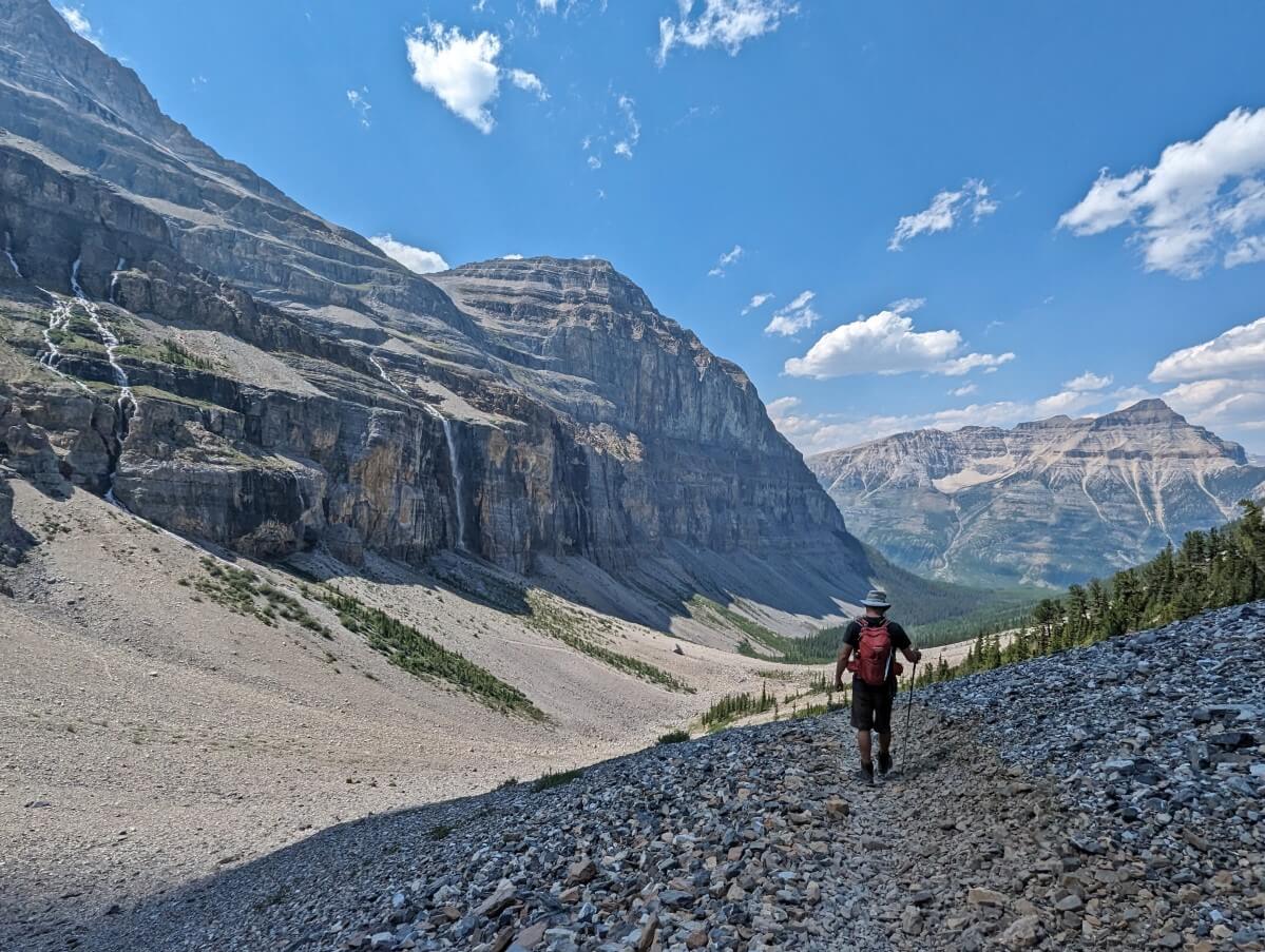 Back view of JR descending steep scree slope on Stanley Glacier Trail extension loop with steep cliffs and waterfalls on left