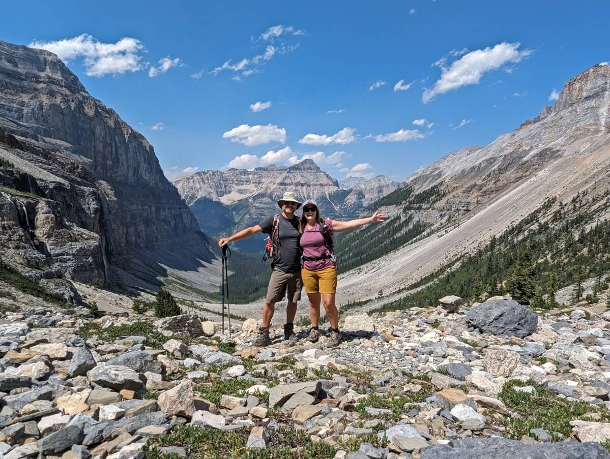 Gemma and JR facing camera with arms out in front of spectacular basin view on Stanley Glacier Trail, with tall mountains all around