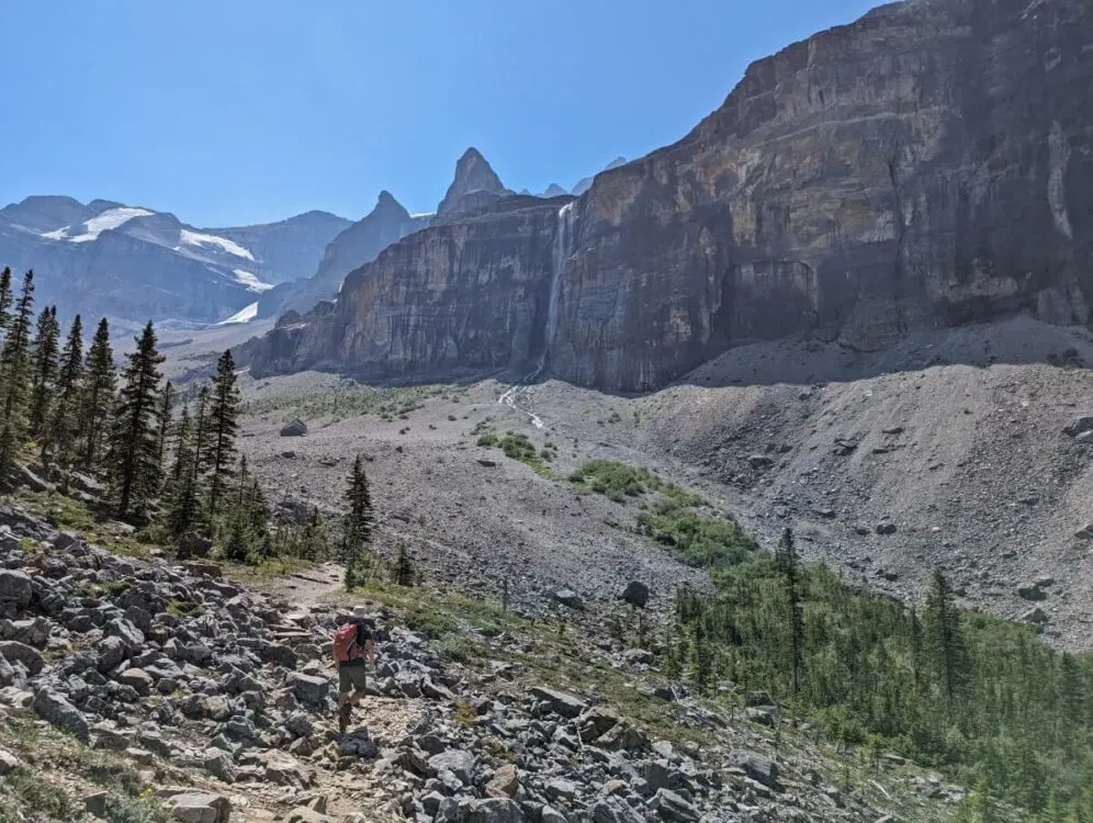 Back view of JR hiking through rocky path through mountainous basin area with waterfall and glacier in background