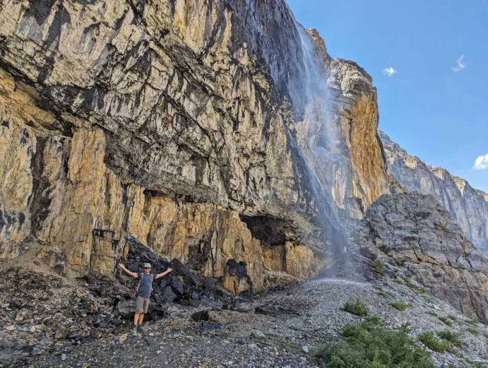JR standing beneath large cascading waterfall, arms outstretched, with steep cliffs on left