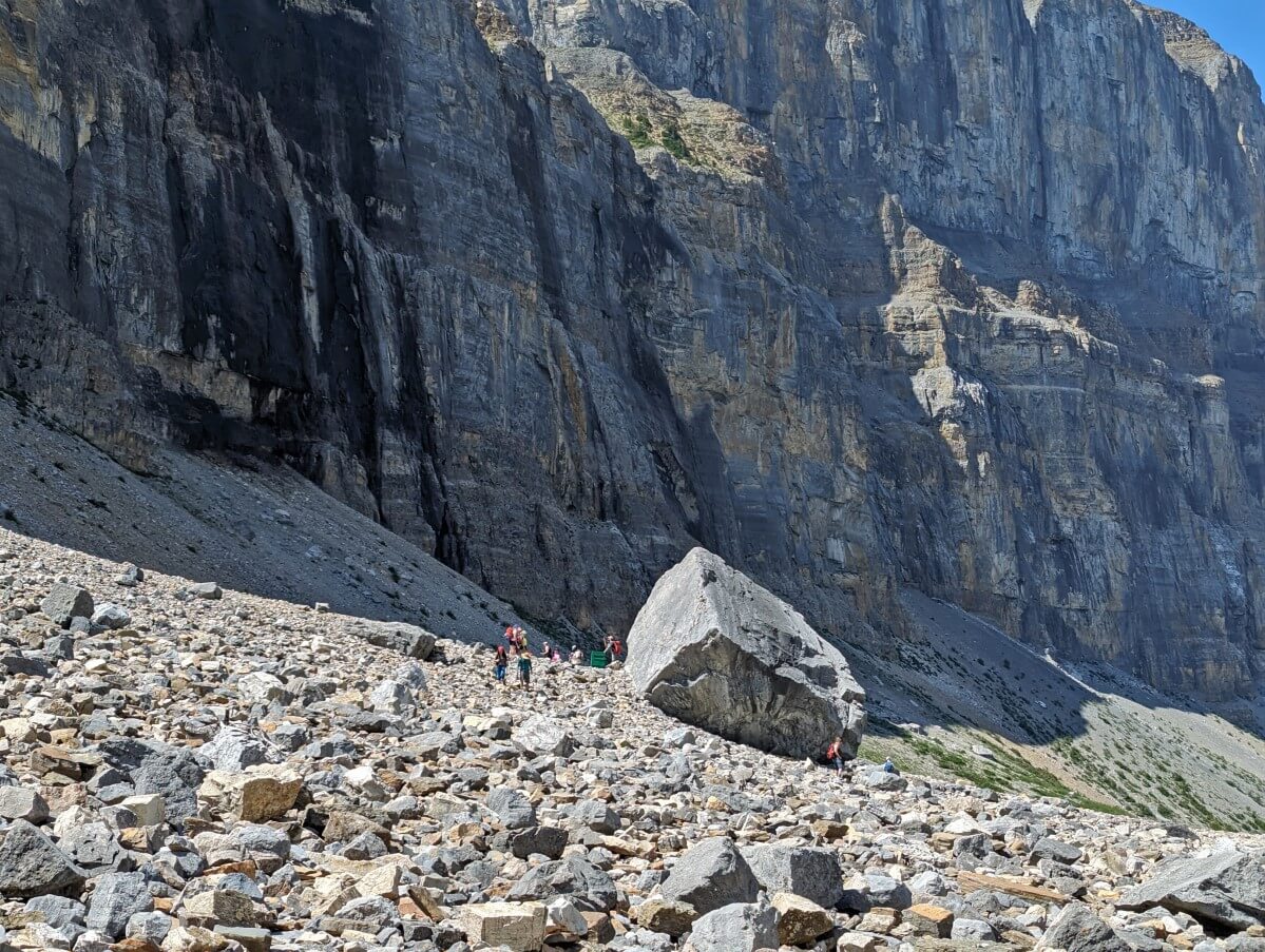 Huge boulder in the middle of boulderfield with group of people searching in rocks below. Steep cliffs behind