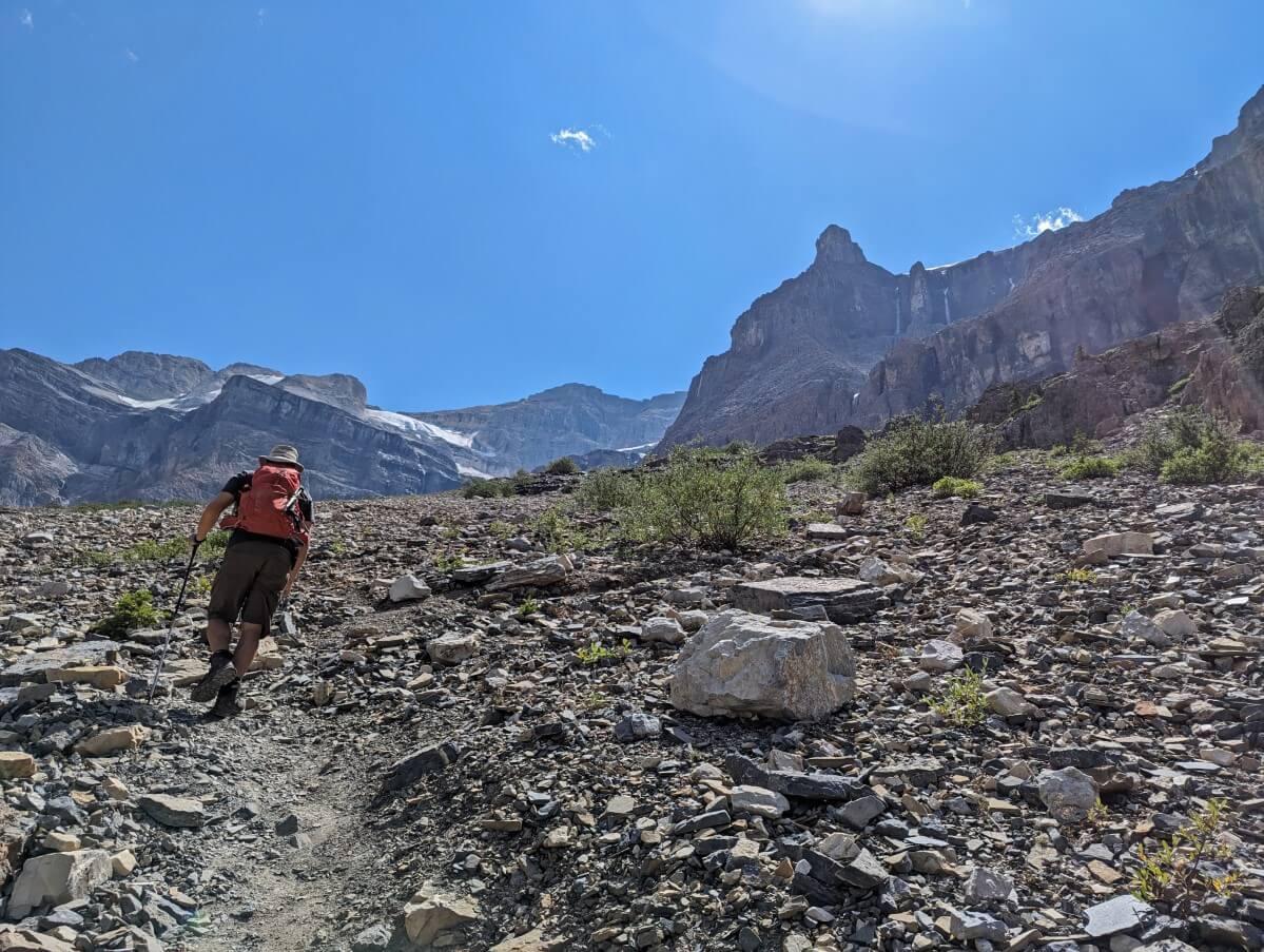 Back view of JR hiking up very steep section of scree path on Stanley Glacier Trail, with mountain peaks behind