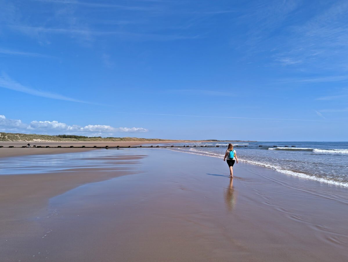 Back view of Gemma walking barefoot on wet sand on Druridge Bay Beach on the Northumberland Coast Path, with reflection in water below