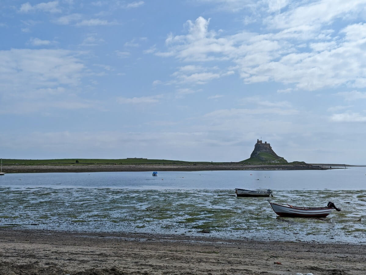 Looking across muddy low tide beach to calm bay with hiltop Lindisfarne castle in background. Two empty boats in foreground