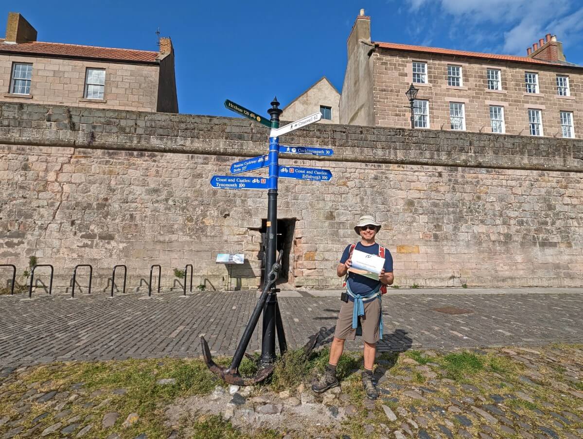 JR standing holding Northumberland Coast Path completion certificate next to anchor with trail signage, in front of brick wall