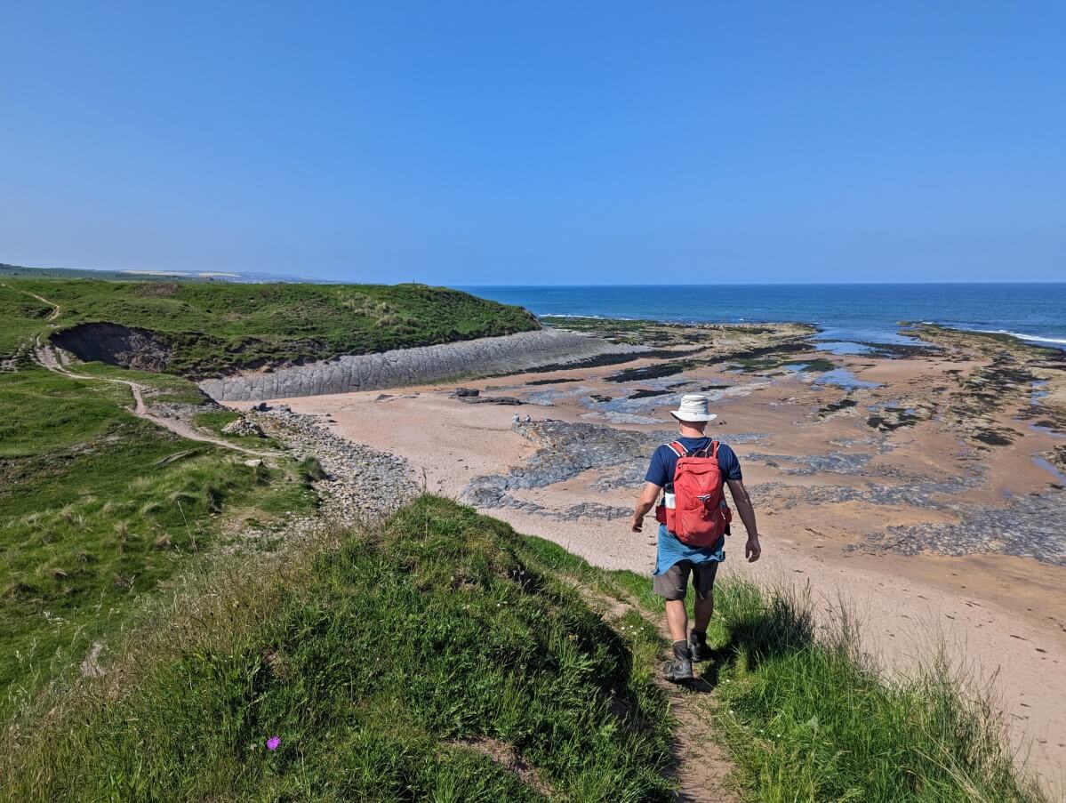 Back view of JR walking on dirt Northumberland Coast Trail trail with sandy beach and calm North Sea on right and sloped grass hill on left