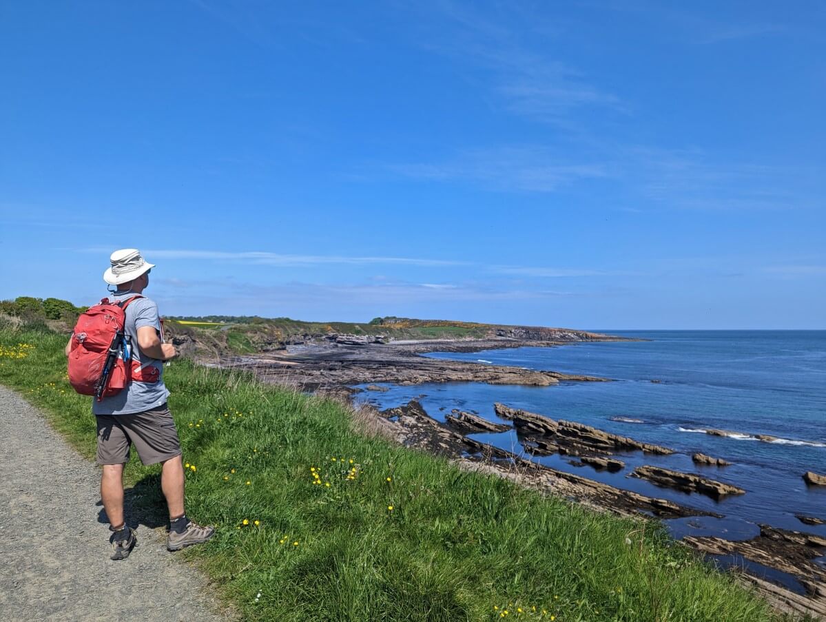 Back view of JR standing on path looking out to calm North Sea on sunny day while walking the Northumberland Coast Path near Craster. The shoreline is rocky