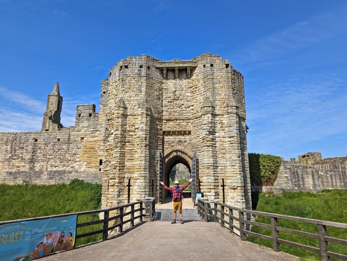 JR stands with arms oustretched in front of medieval gate at Warkworth Castle on the Northumberland Coast Path