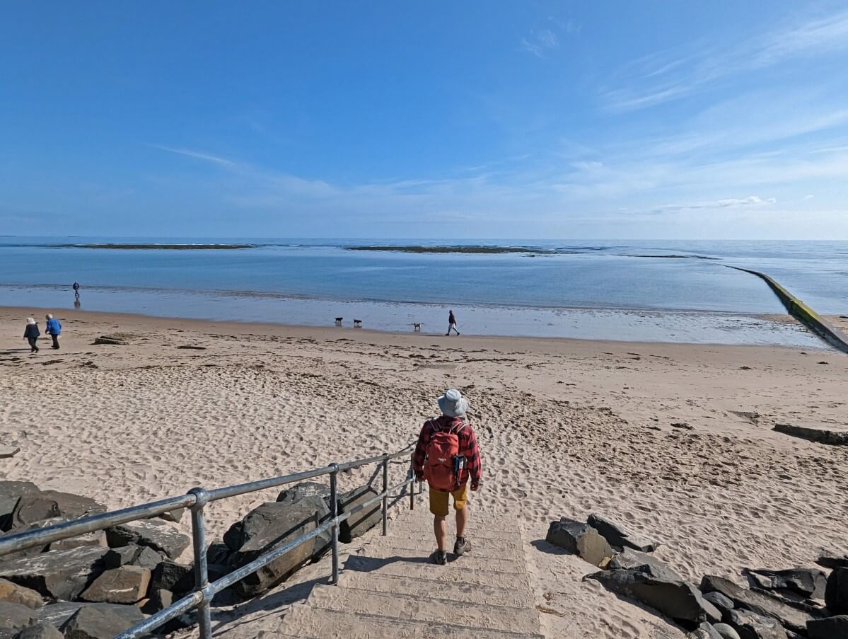 Back view of JR walking down concrete stairs to sandy Cresswell Beach in Druridge Bay. There are a handful of people walking on the beach